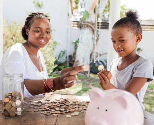 African mother in her back yard counting coins with her daughter and throwing them into a piggy bank.